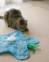 A tabby cat crouched on a rug stalking the fish-print play mat, the white chirping unit in the centre and the green feather lure poking out at the edge